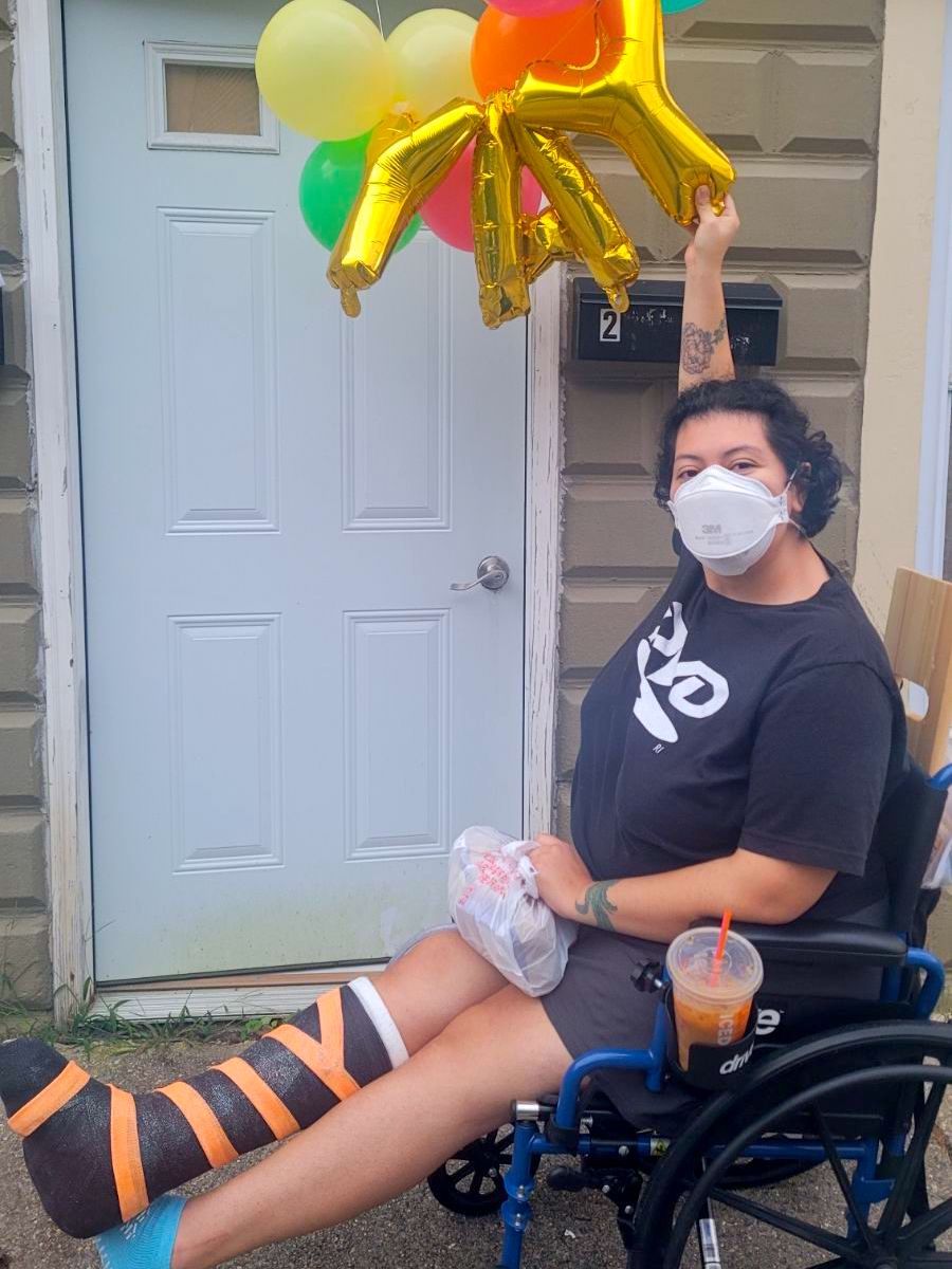 Myself, an olive-toned individual with short hair wearing an aura mask sitting in a wheelchair with a black and orange cast. My wheelchair is in front of the door to my apartment and I'm holding gold balloons that spell out YAY